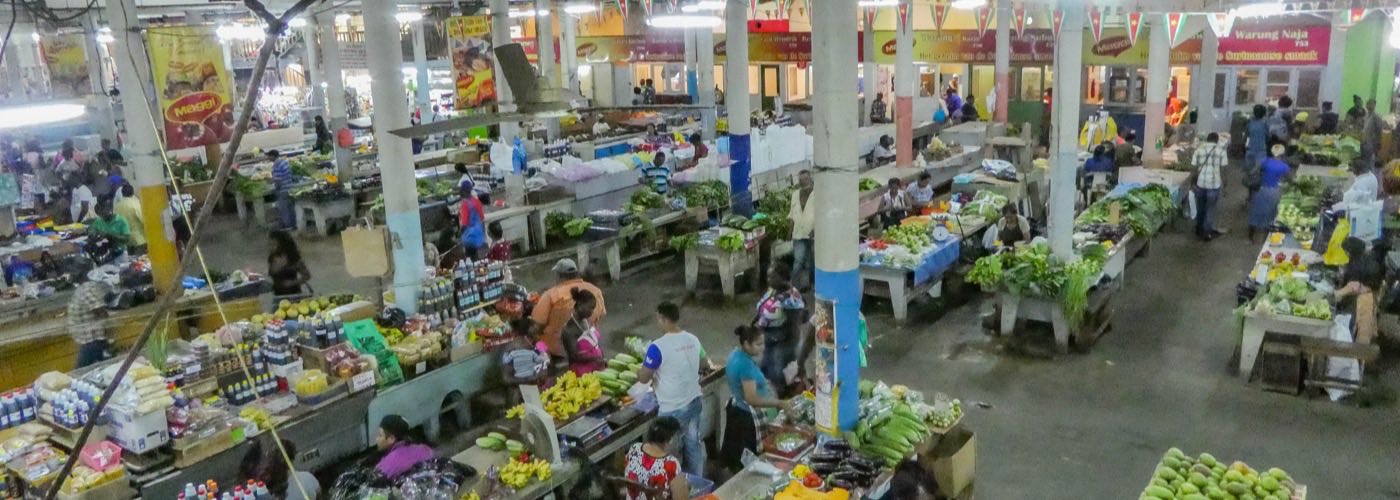 Stalls of fruit and vegetables under the concrete roof of the Centrale Markt in Paramaribo, with shoppers walking between them.