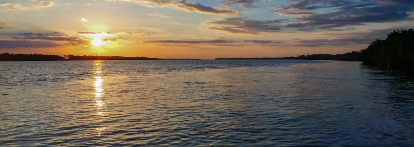 The sun setting over the wide, flat water of the Commewijne River, with low forest along the far bank.