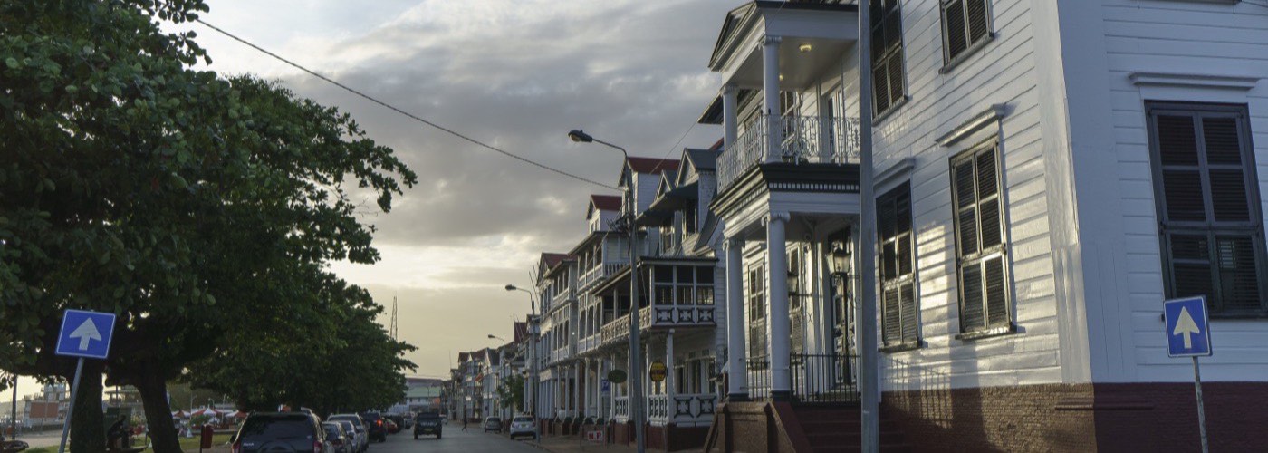 A row of white timber houses with shuttered windows and first-floor verandas along a Paramaribo street at dusk.